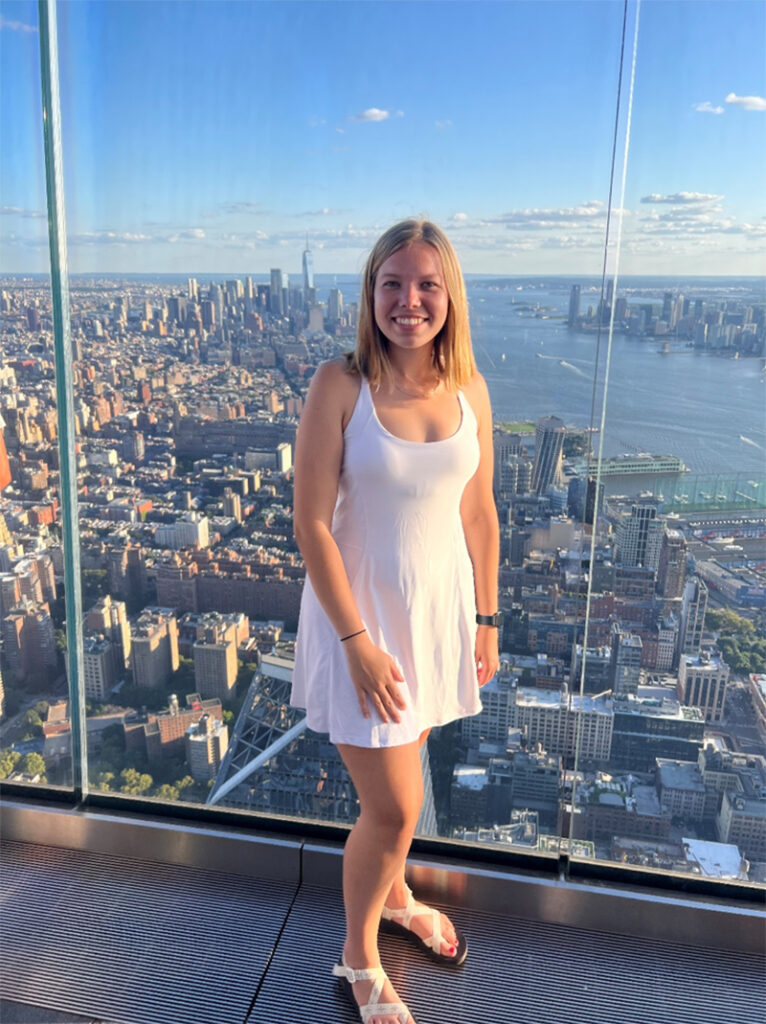 Savannah Tabor poses for a photo atop a skyscraper deck with a view of New York City behind her.