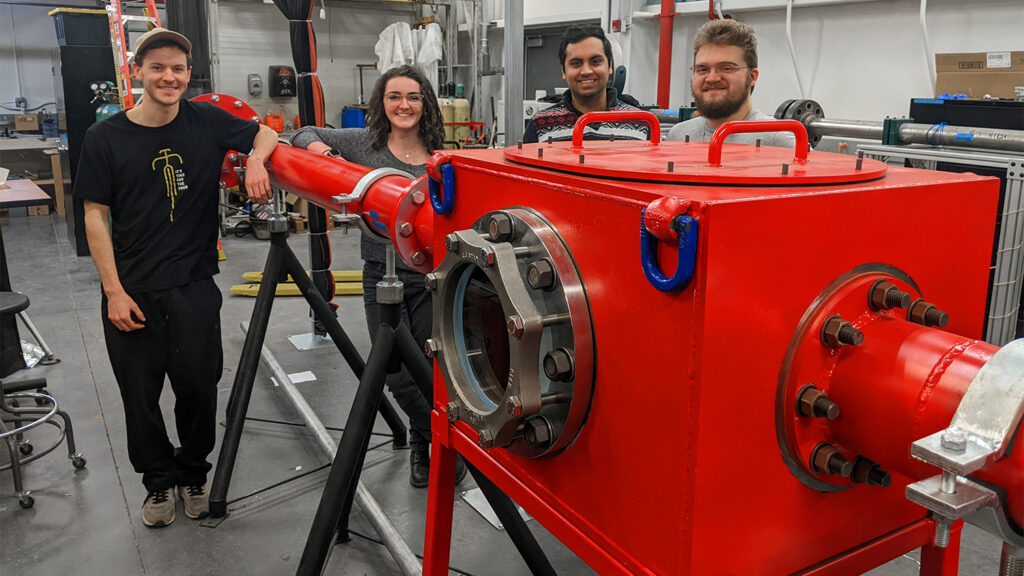 Engineering students stand around the new hypersonic tunnel. The tunnel test box and piping are painted in NC State red and is located in an industrial setting.