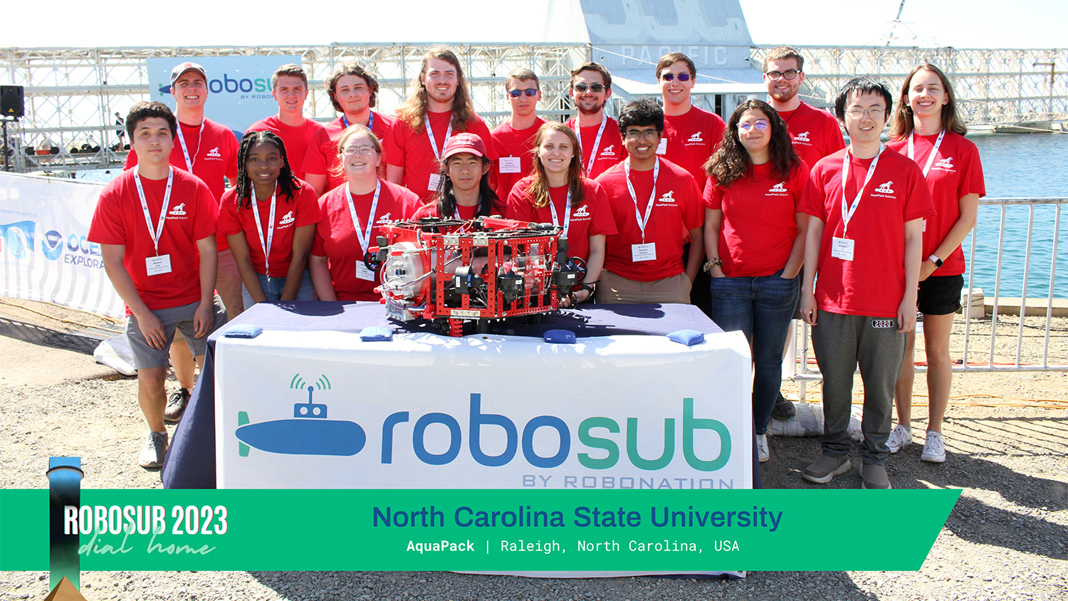 The NC State AquaPack Robosub student-led team poses for a group photo on a sunny day behind their submersible robot which is sitting on a table with a white tablecloth and the Robosub logo in blue and teal on the front.
