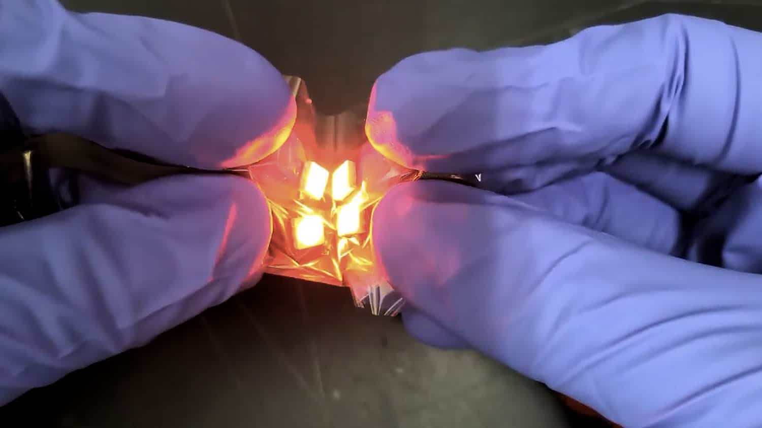 a flexible circuit has been printed onto a piece of polymer. the polymer and circuit are being crumpled by a pair of gloved hands. the circuit is lighting up an LED. the image is a photograph, not an illustration.