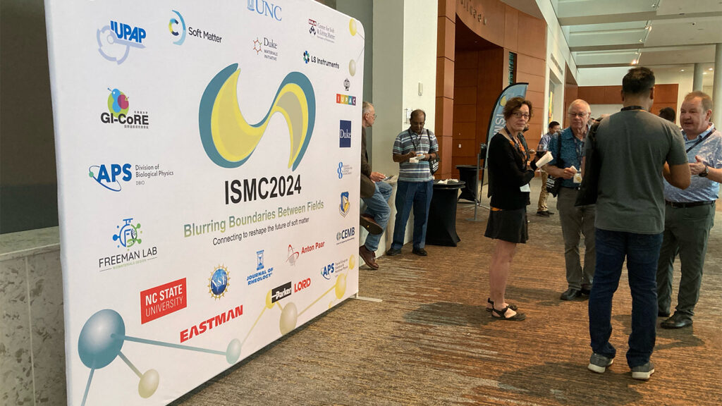 Photo from the International Soft Matter Conference 2024 showing a large banner with several logos of conference sponsors on the left and a group of attendees on the right.