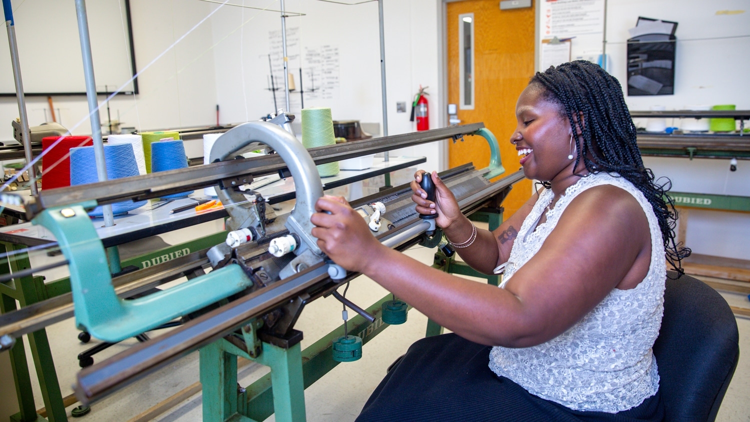 A woman with braided hair is operating a knitting machine in a workshop. She is focused on her work and smiling. Spools of colorful thread are visible on the machine, and the background shows workshop equipment and a door with a safety sign.