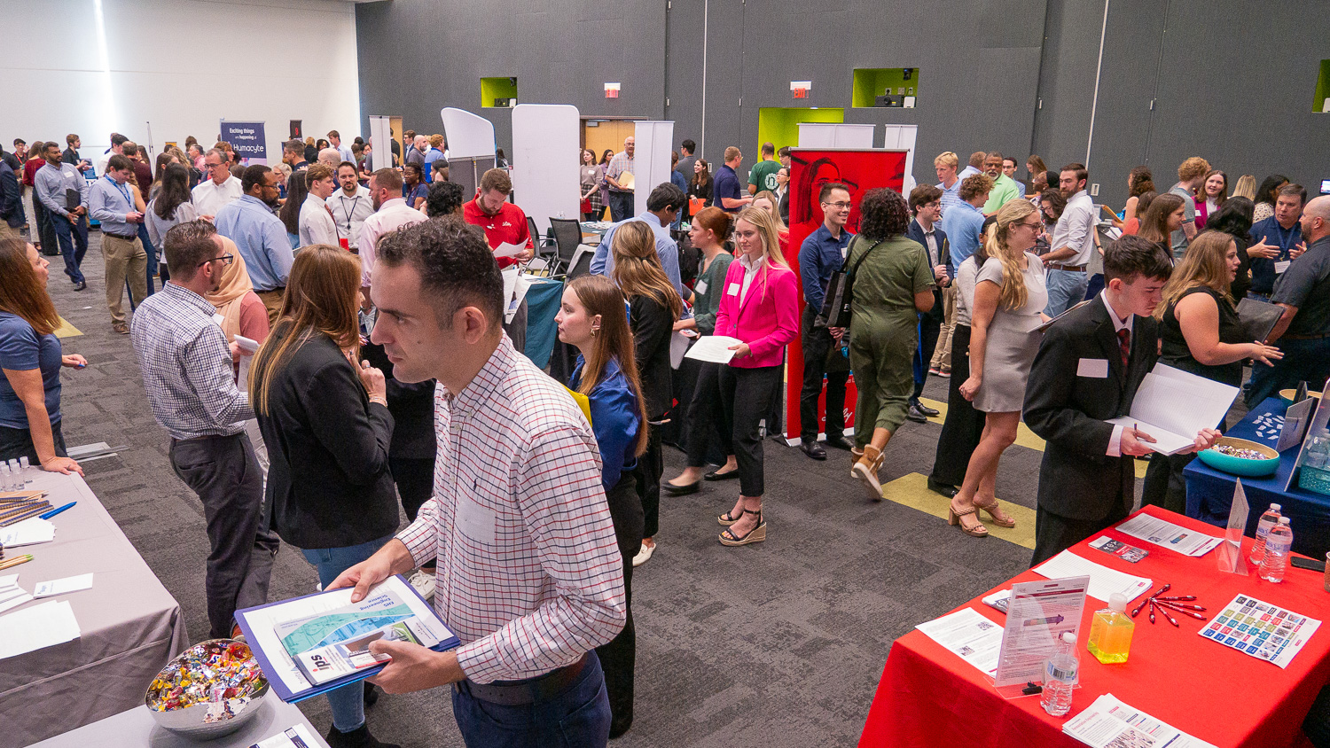 Students mingle and speak with employer representatives stationed at display tables in a large conference hall at Hunt Library on NC State's Centennial Campus. The room has dark gray walls and dark gray carpet.