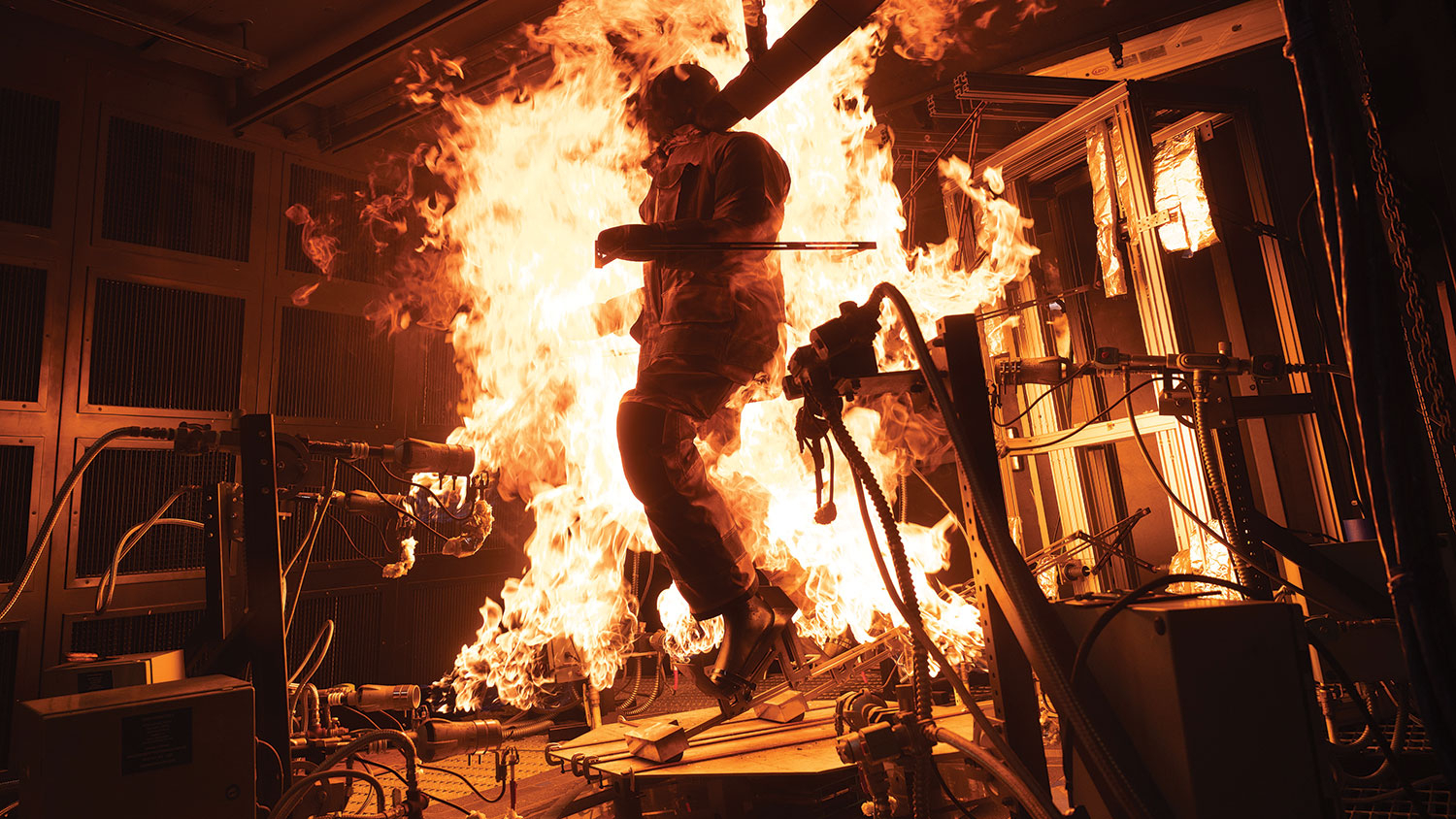 Photo of a manikin dressed in a fire-protective suit that resembles heavyweight coveralls. The whole suit is engulfed in flames, which are being shot at it from all directions in a science lab.