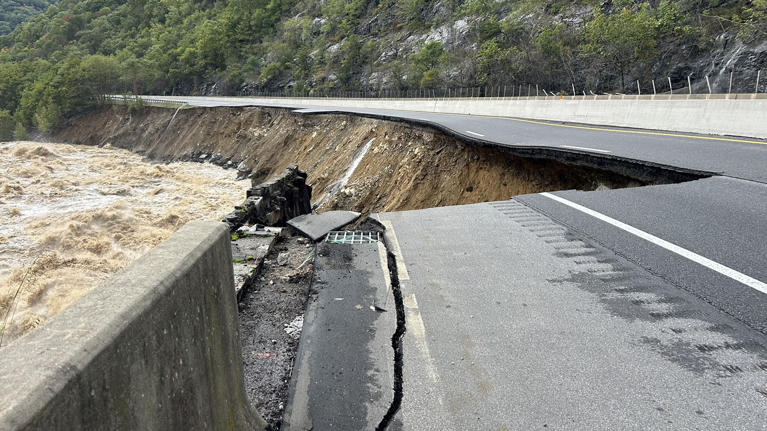 a roaring river rushes by a road. half of the road has been washed away by the river