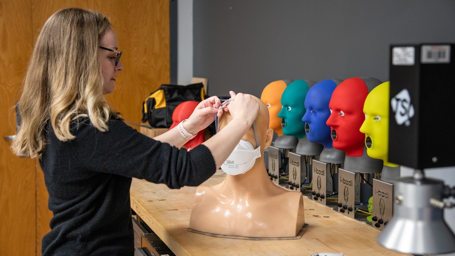 Melissa Armistead adjusts a mask on a mannequin head. Several colorful mannequin heads are lined up on a workbench, each with an open mouth, which is part of a research or testing setup. A door and equipment are visible in the background.