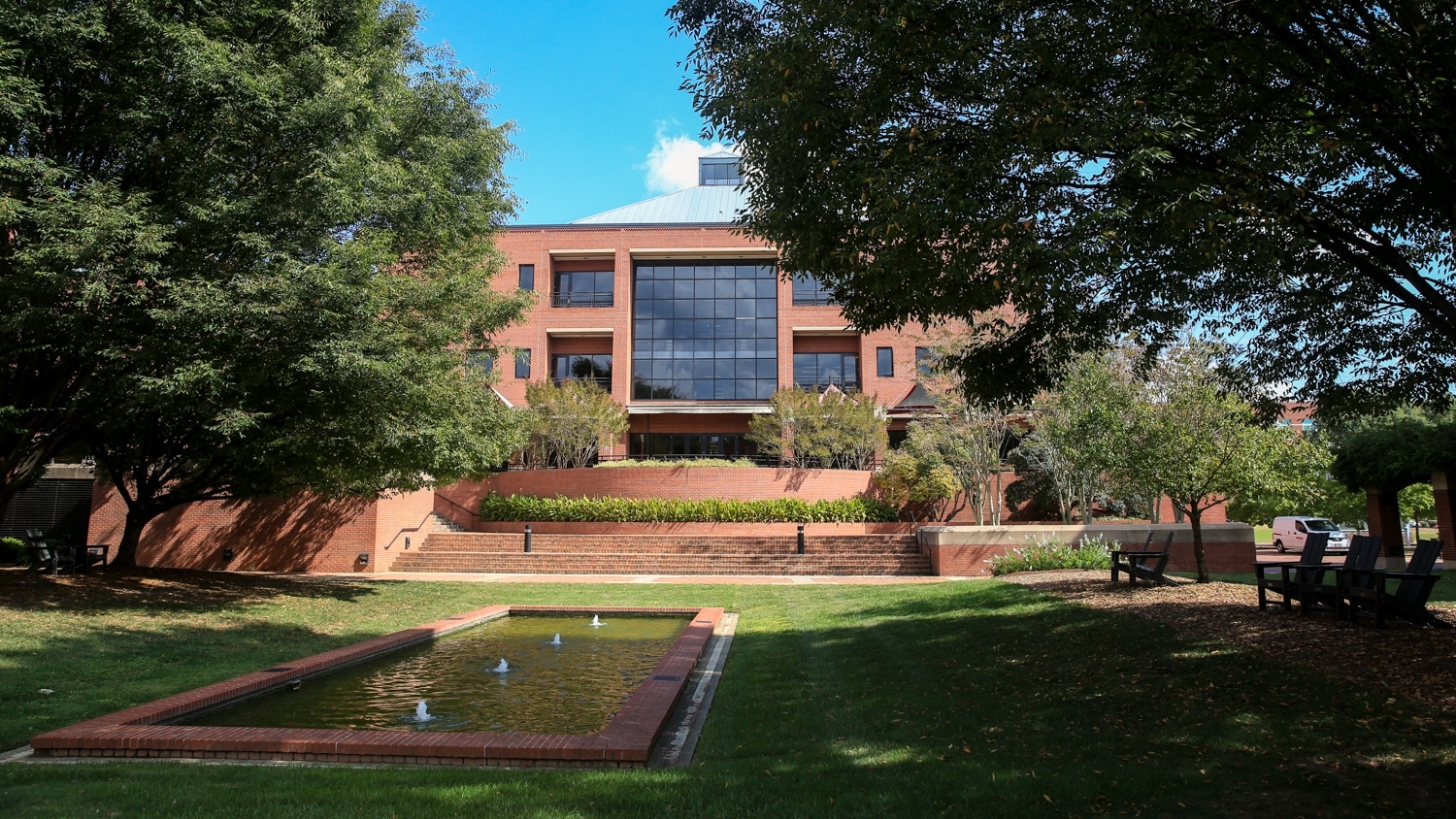 A serene outdoor courtyard features a rectangular reflecting pool surrounded by lush green grass and trees. In the background, a modern brick building with large reflective windows (the Wilson College of Textiles) stands tall under a clear blue sky. Adirondack chairs are scattered around the area.