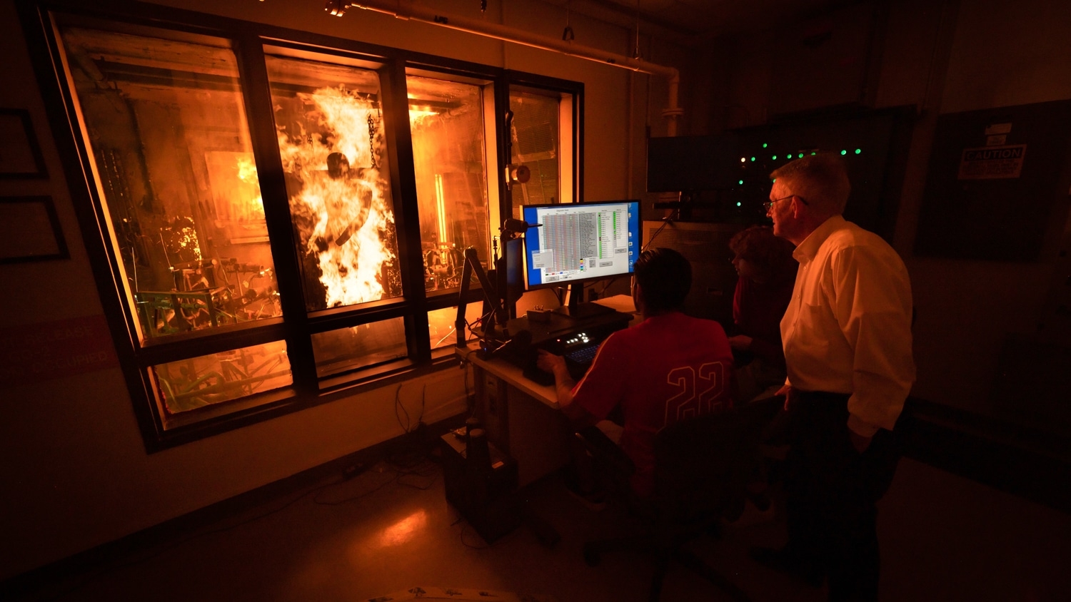 A group of people monitors a Dynamic PyroMan test in a TPACC laboratory. One person is seated at a computer workstation with data on the screen, Roger Barker and others stand nearby. A large blaze is visible through a window, contained in a separate room.