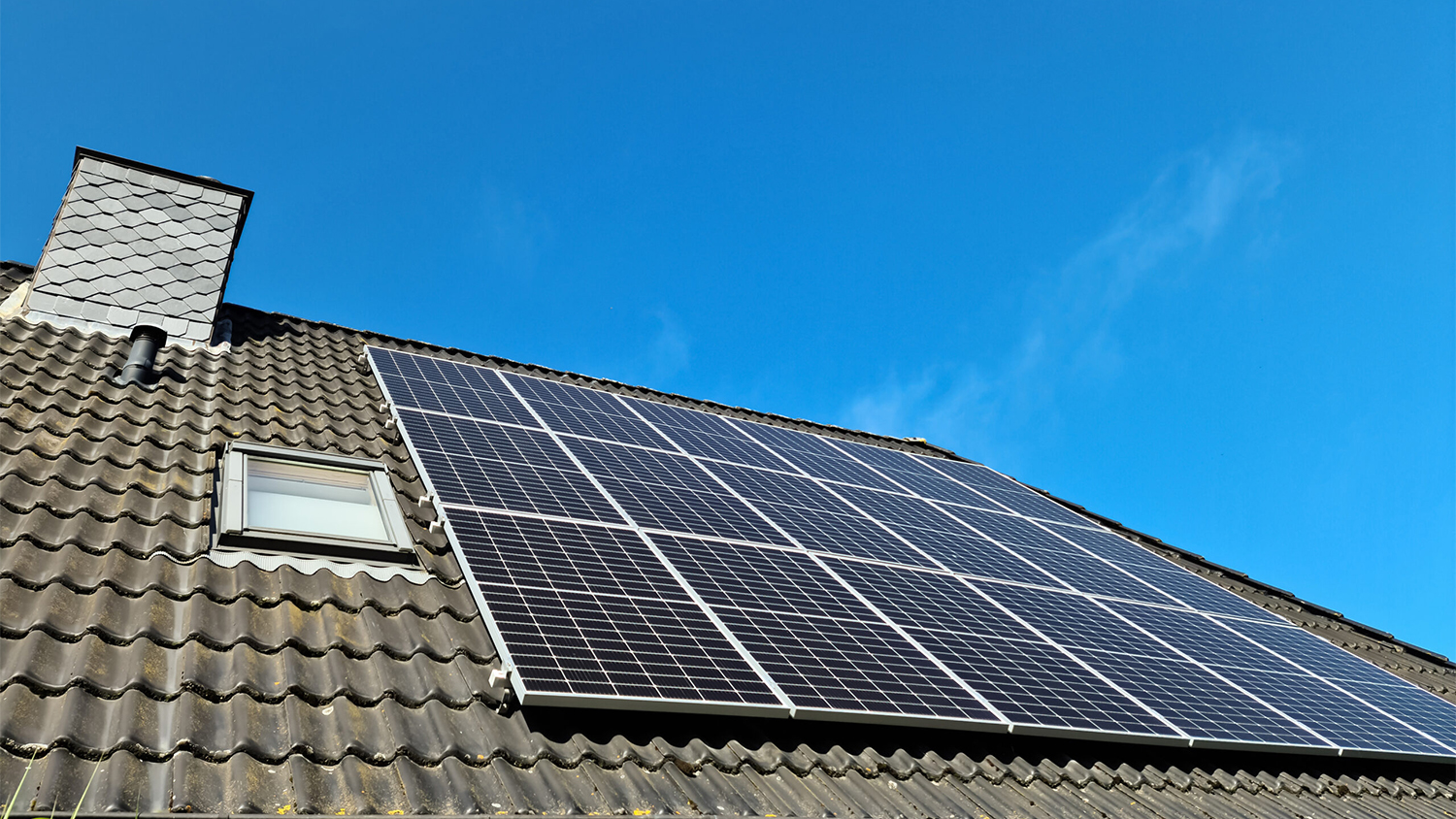 A series of black solar panels on a residential roof; a chimney is off to the left and the sky if a deep blue with a wisp of white clouds.