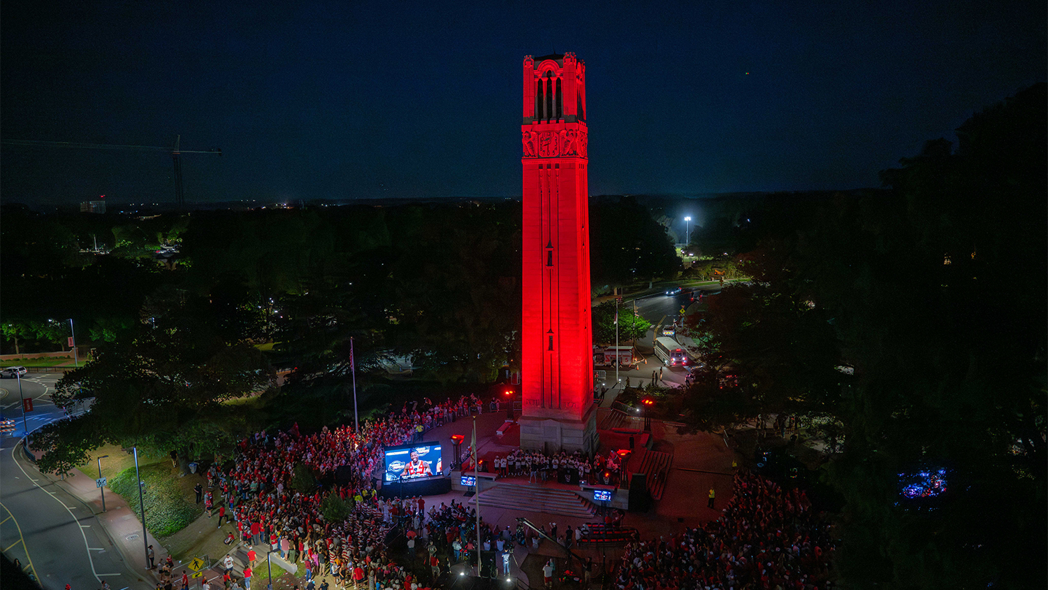 NC State basketball fans celebrate the return of the Men's and Women's 2023-2024 final four teams at a celebration at the Belltower. It is night and the belltower is lit in NC State red uplighting. Downtown Raleigh can be faintly seen in the background.