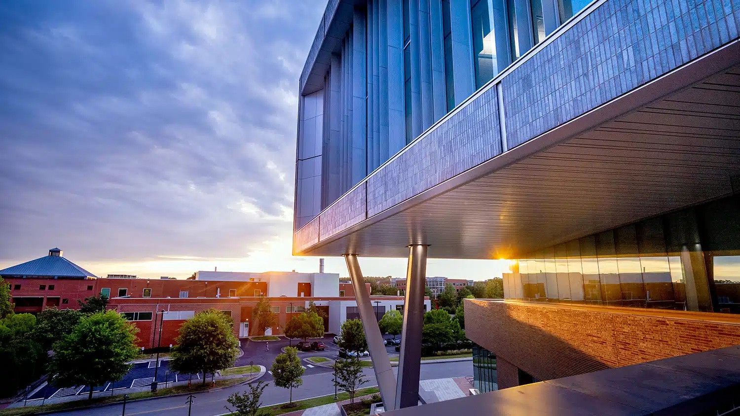 The sun sets behind a corner of Fitts-Woolard Hall. the sky is partly cloudy and bluish pink.