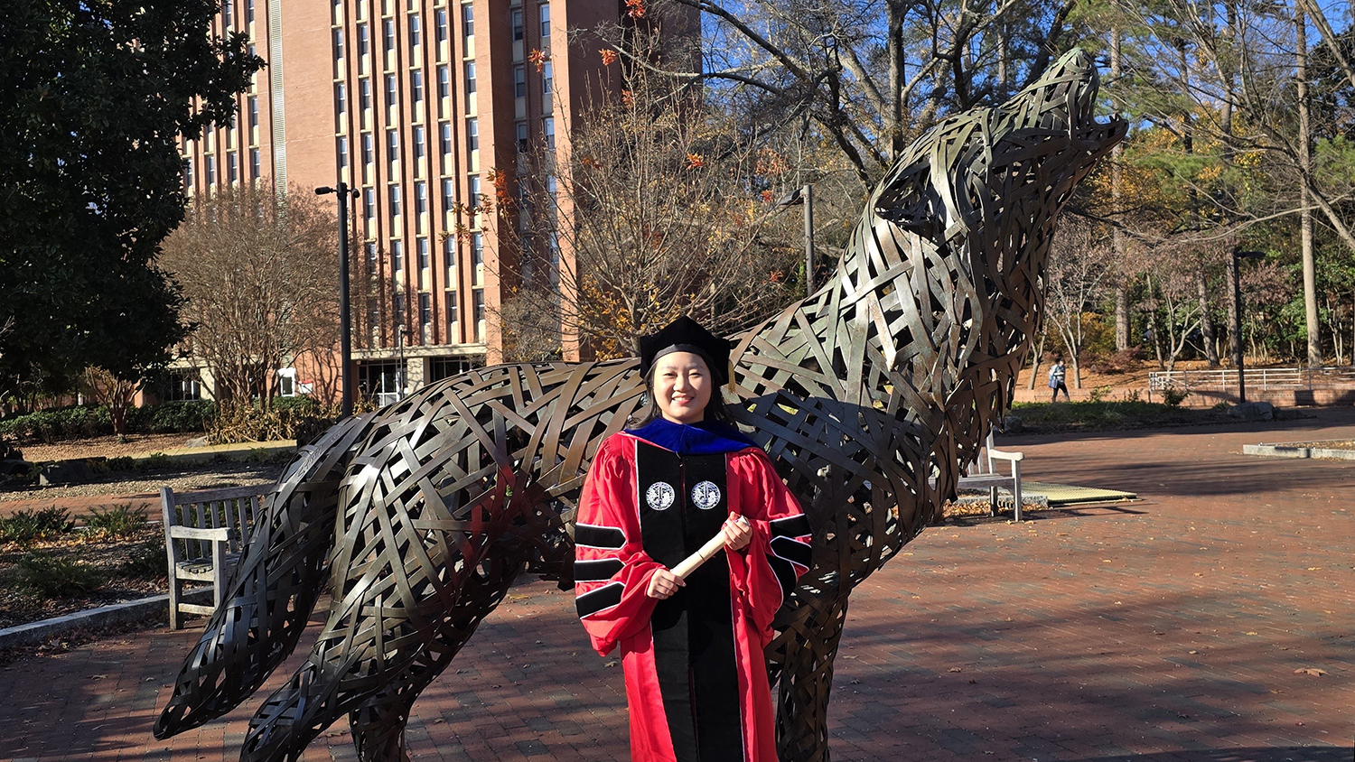 Emily Duan wearing graduation cap and gown while standing next to wolf sculpture at Wolf Plaza on NC State's campus.