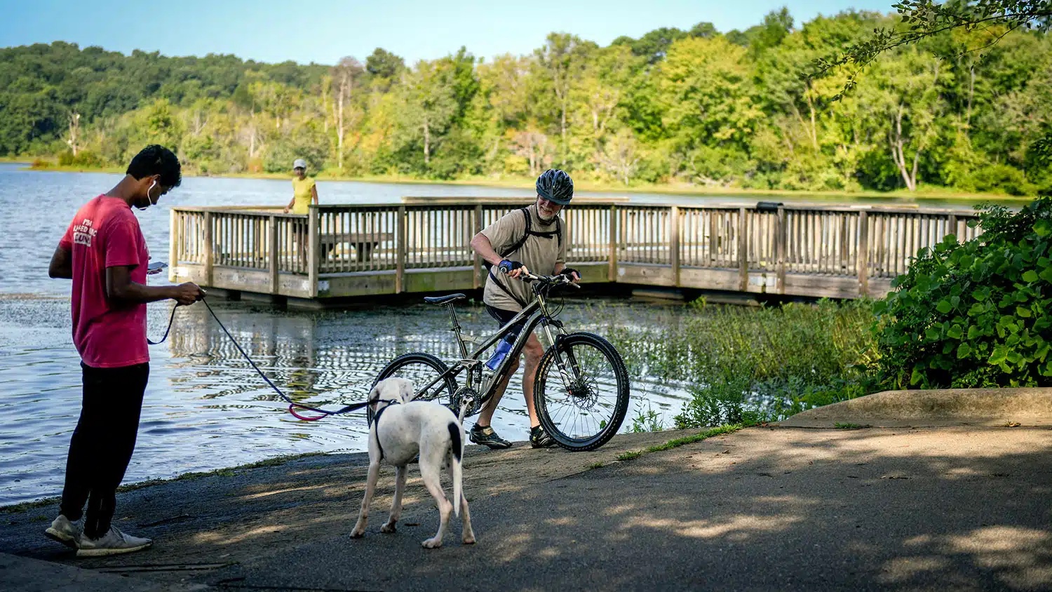 Centennial Campus amenities include Lake Raleigh's waterfront. At the shoreline, a man on a bike smiles at another man walking their dog. A woman wearing a yellow top is in the background standing on a wooden pier jutting out into the water.