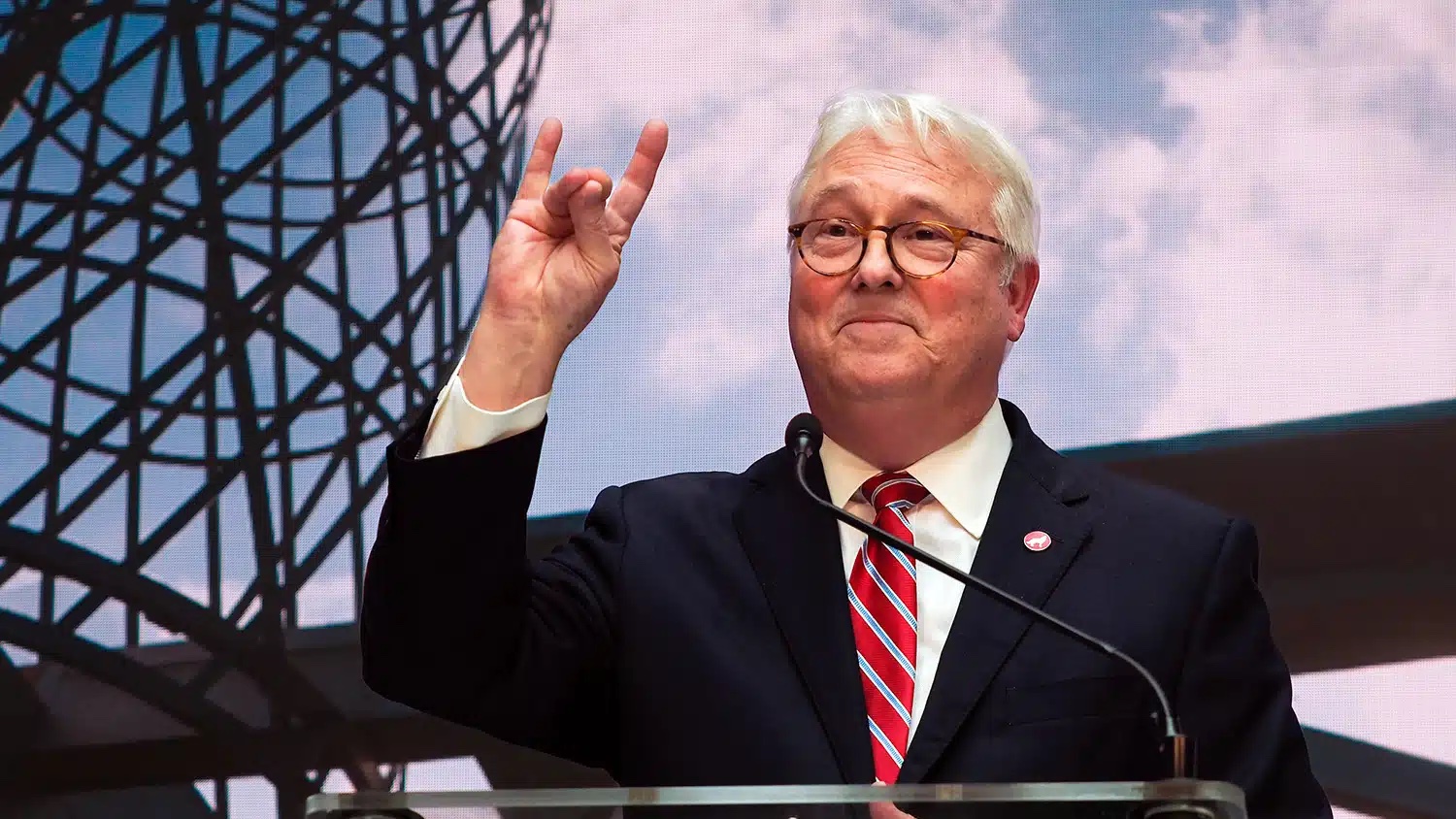 Chancellor Randy Woodson flashes the NC State wolf hand sign while standing in front of a large LED screen showing a portion of Talley Student Union.