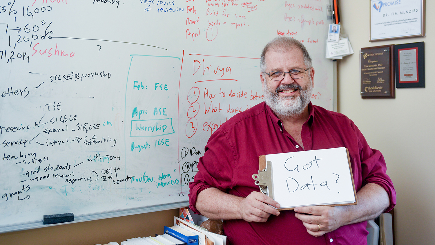 Tim Menzies in his office holding a small whiteboard with "Got Data?" written in black marker on it. He is standing in front of a large wall-sized whiteboard covered in writing.