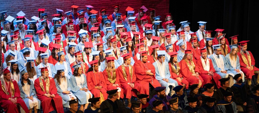 A group of Joint Department of Biomedical Engineering students sit dressed in red gowns from NC State and light blue gowns from UNC during a commencement ceremony.
