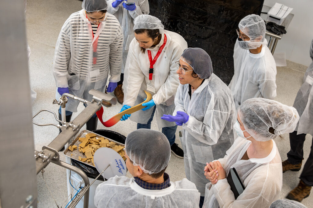 A group of researchers and lab workers dressed in white lab coats and hair nets gather around Bühler extruder containing lab grown protein.
