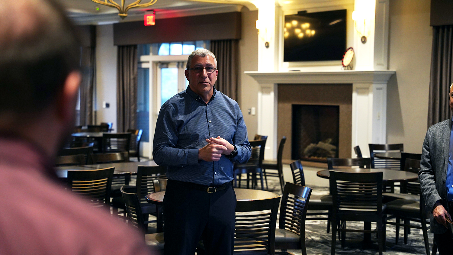 Basil Hassan, center speaks with two individuals framed on the left and right facing him in a traditionally styled dining room with carpet, fireplace and draperies.
