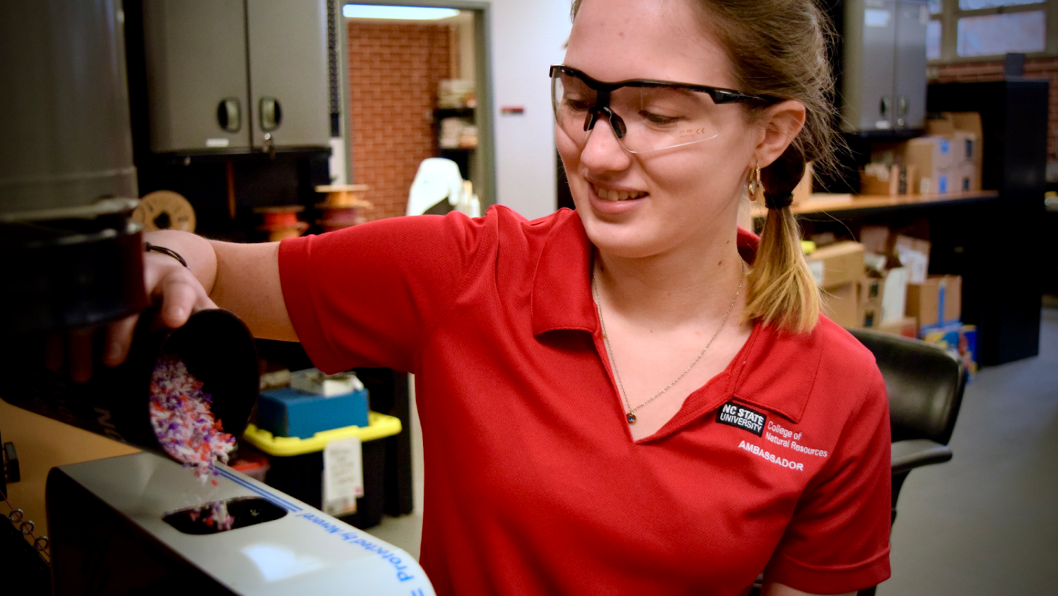 Female student wearing a red NC State polo shirt pours waste into a receptacle.