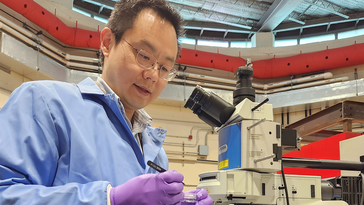 Ge Yang dressed in a blue lab coat and wearing glasses and purple gloves works in his lab.