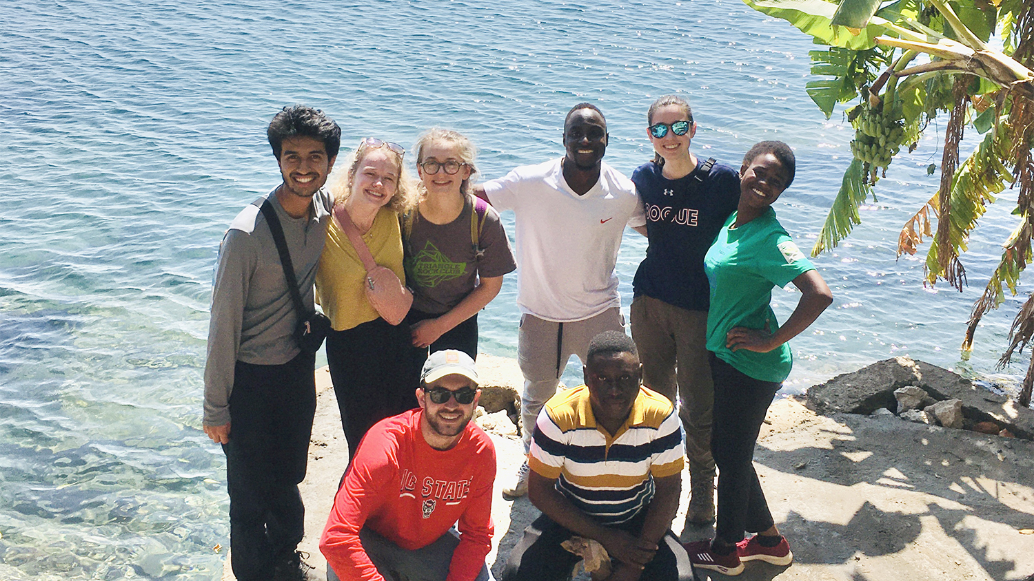 NC State students pose for a group photo on a Malawi beach next to the light blue ocean water behind them and large palm tree leaves on the right.