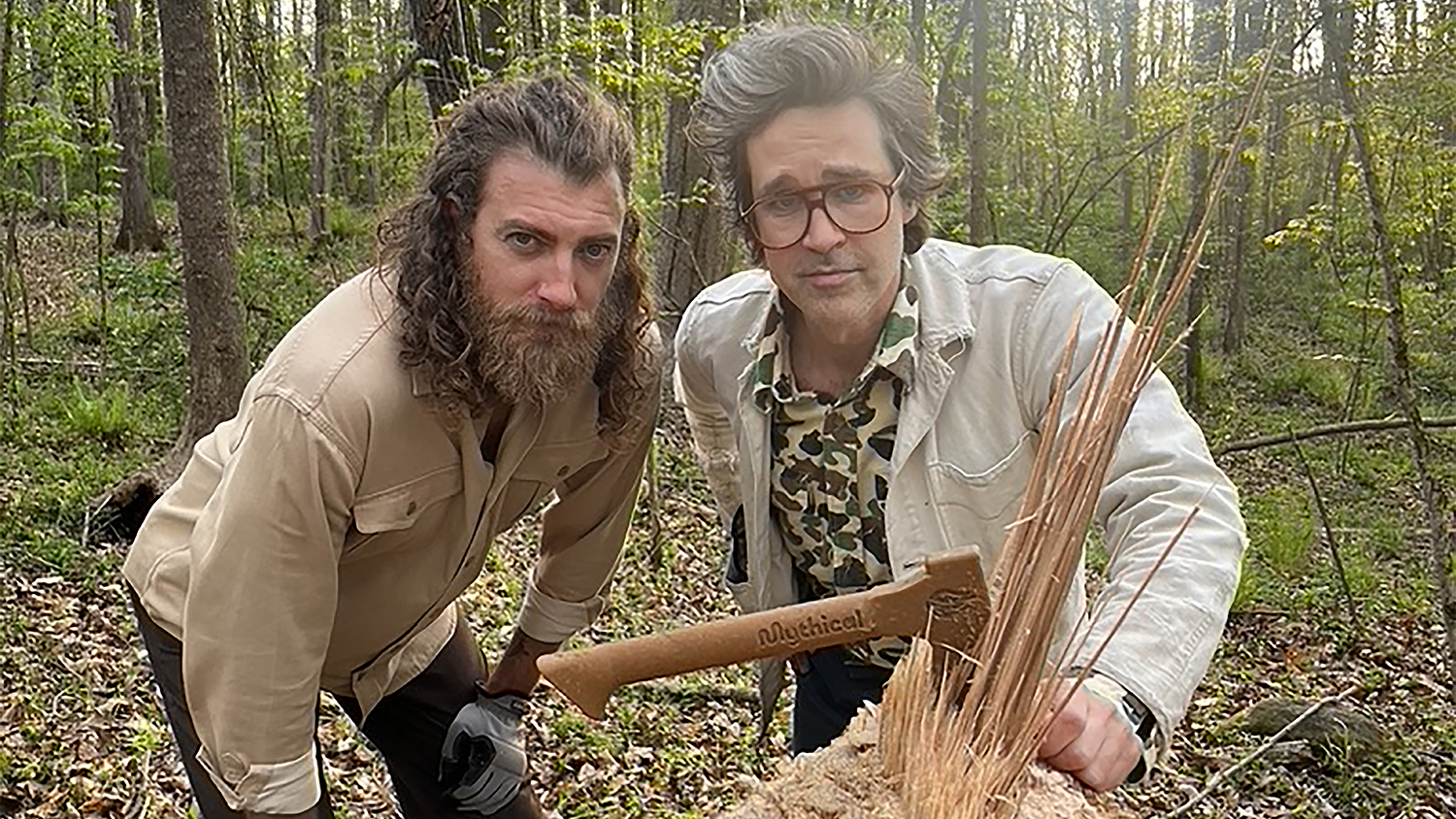 Rhett McLaughlin and Link Neal stand next to a stump from a felled tree with their peanut butter axe embedded in the face of the stump.