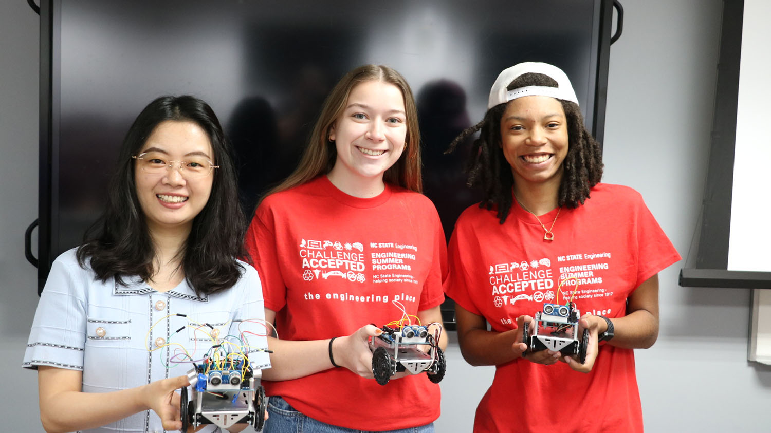 Lirong Xiang (left) with students during the 2024 high school robotics summer camp. All three are holding small robotic devices. Lirong is wearing a light blue top and the two to her left are wearing red t-shirts.