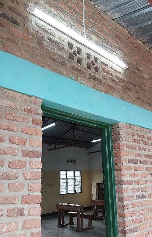 image shows a strip of outdoor lighting above the doorway of a brick building; a horizontal beam painted in teal crosses the side of the building above the doorway.