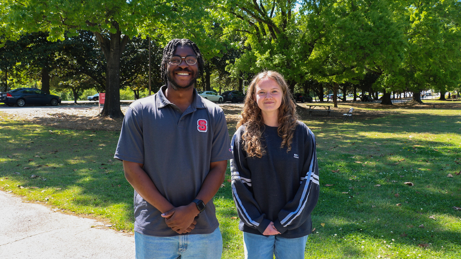Two BAE students pose for a photo while standing on a sidewalk next to a green lawn and trees in the background on a sunny day.