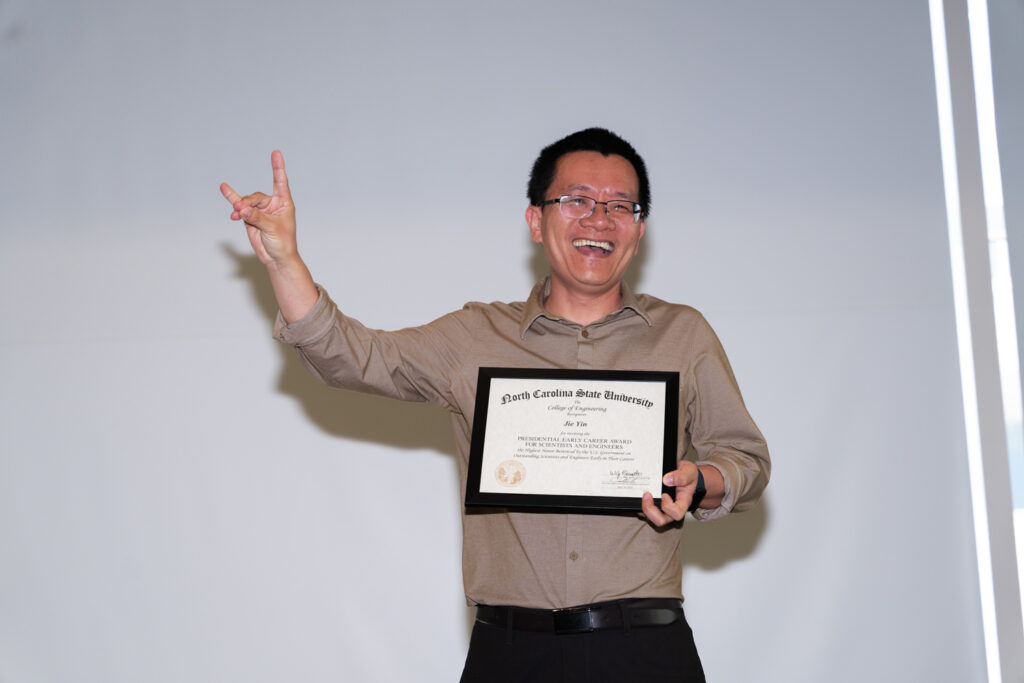 Jie Yin laughs and shows the NC State wolf sign after receiving his framed award which he holds in front of him.