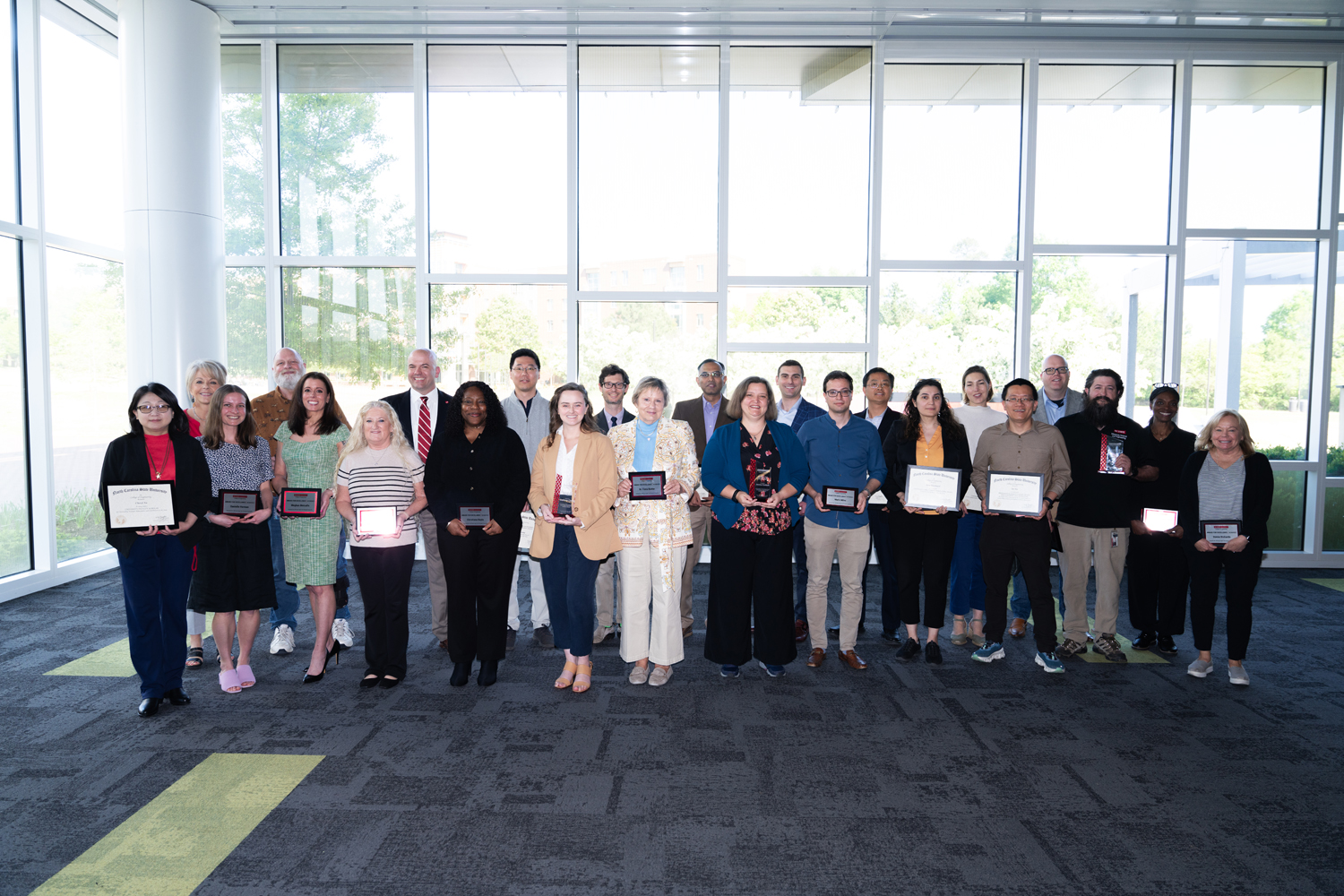 Group of faculty and staff pose for a group photo in front of a large wall of windows during an awards ceremony.