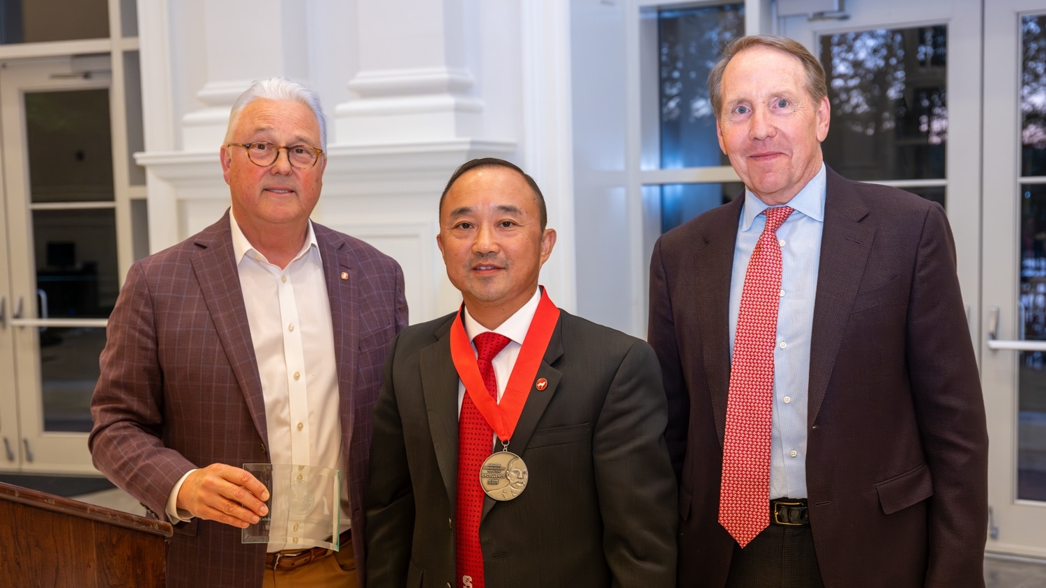 Three men in suits pose indoors: From left to right: Randy Woodson, Xiangwu Zhang and Ed Weisiger Jr. They stand in front of white walls and glass doors, with a podium to the left. Xiangwu Zhang wears the Holladay Medal. The lighting is bright and the atmosphere formal.