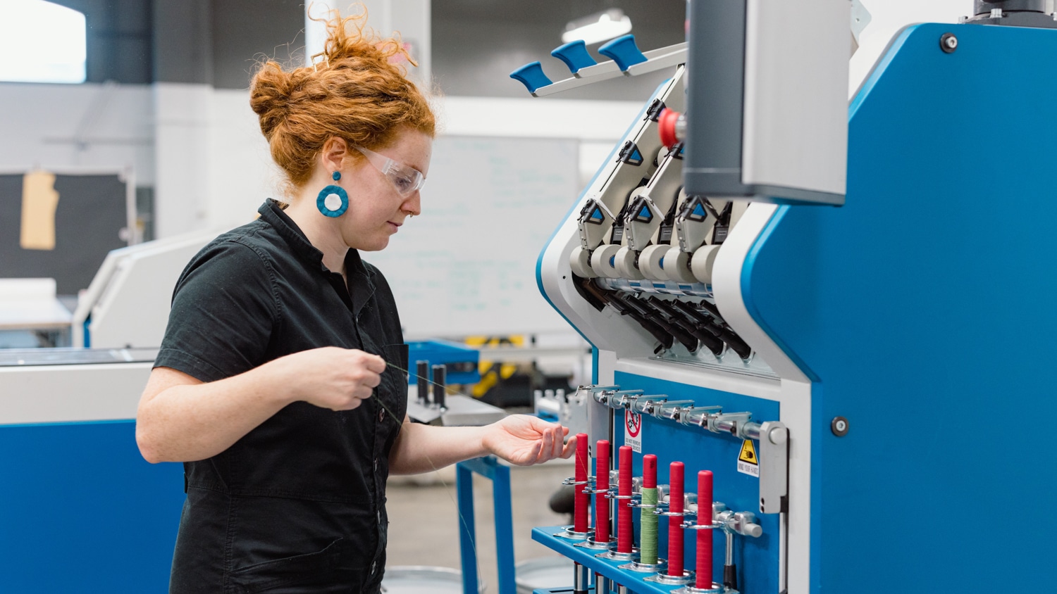Bailey Knight operates a blue industrial machine in the Wilson College of Textiles' Flex Factory. She wears large blue earrings and a black shirt. Spools of red thread are visible on the machine, indicating a textile or sewing process.