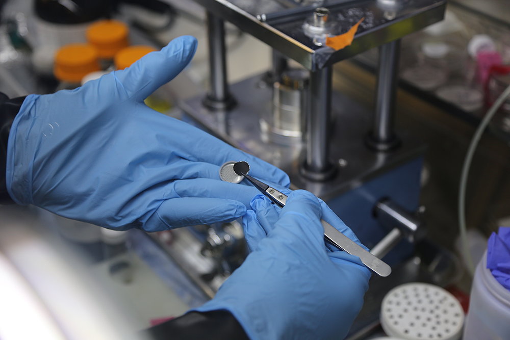 Blue latex gloved hands hold a pair of tweezers in the right and half of a CR2032 battery in the left.