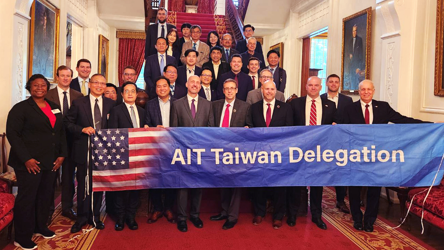 The delegation and representatives from NC State at the North Carolina Executive Mansion. They are grouped together on a large grand staircase for a group photo. The walls are ivory and the carpet is red. The front row holds a banner reading "AIT Taiwan Delegation" in white letters on a blue background.