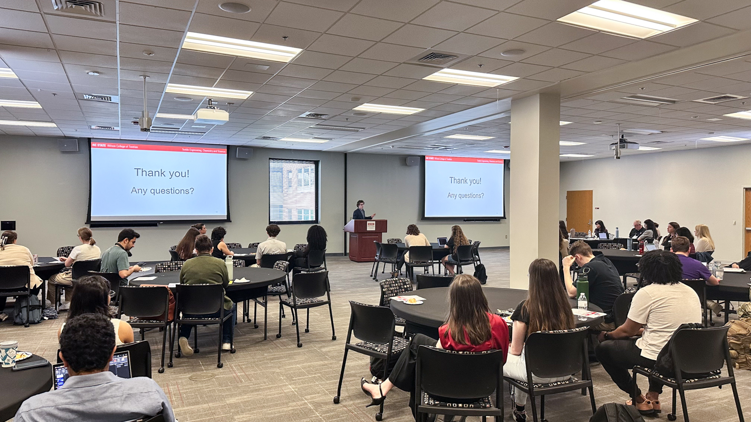 A speaker presents at a podium in a conference room with several round tables of seated attendees. Two screens display the message, "Thank you! Any questions?"