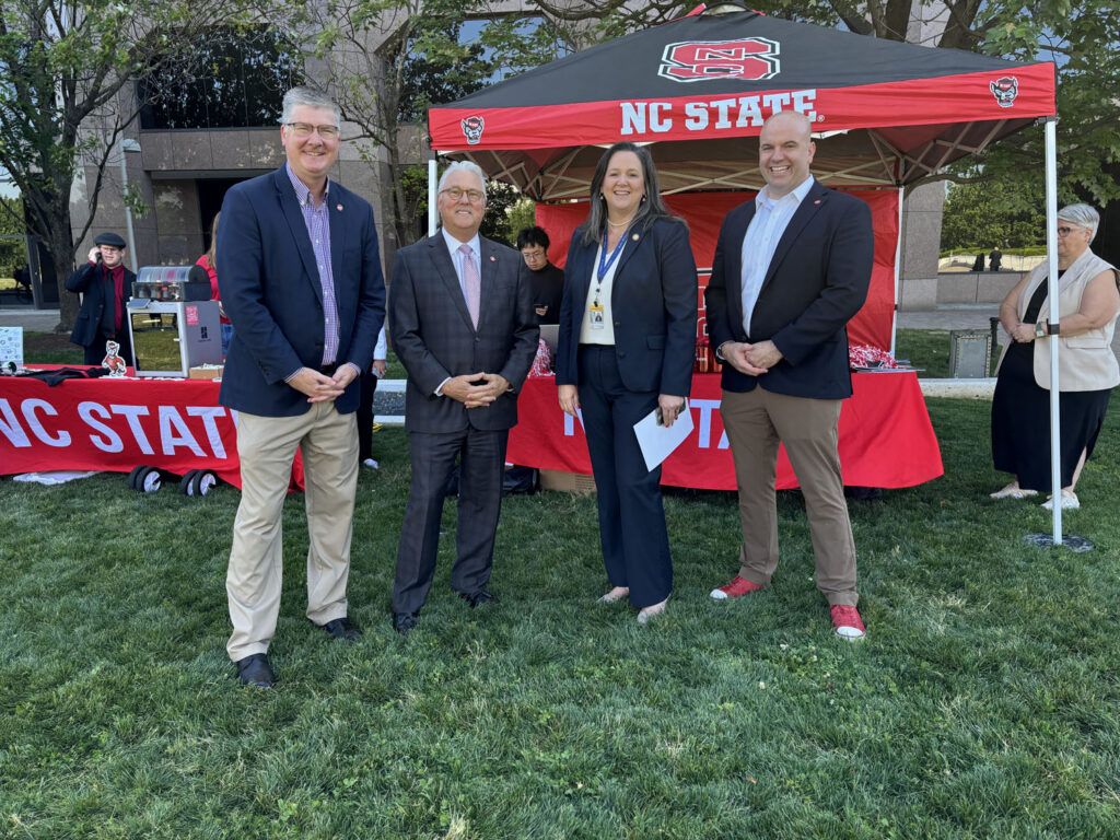 CALS dean Garey Fox, outgoing NC State chancellor Randy Woodson, an unidentified woman, and COE dean Jim Pfaendtner pose for a photo in front of a NC State table and canopy decorated in red, black and NC State logo.