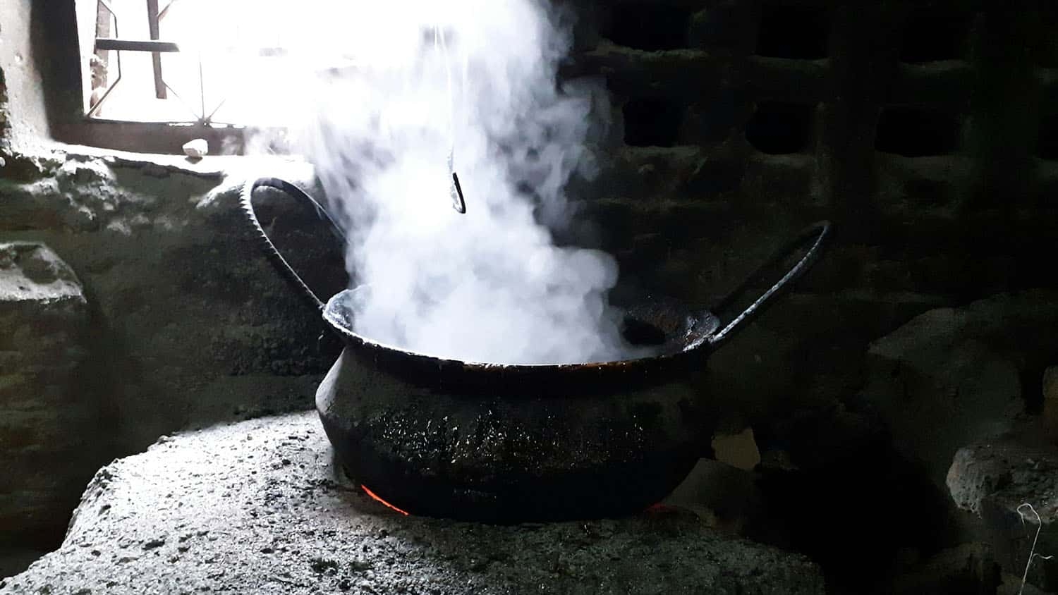 steam billows out of a black metal pot that rests on a stone oven. there is an open fire in the oven.