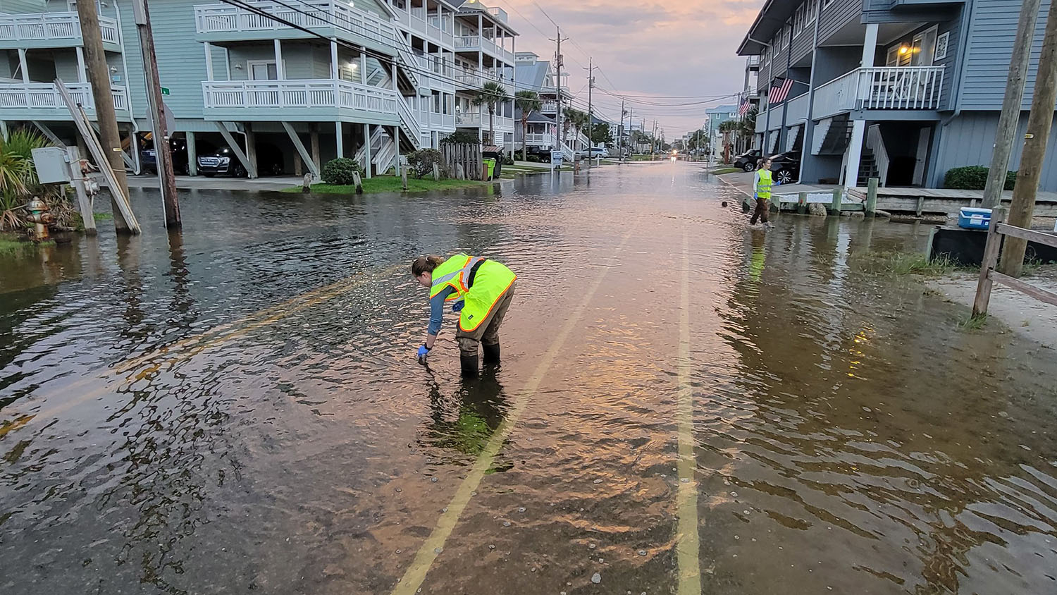 a person wearing a bright yellow vest fills a sampling jar with water from a flooded street