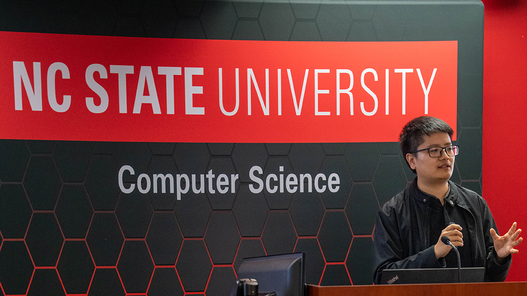Yuchen Liu, holding a microphone while standing behind a lecturn, addresses a seated audience.