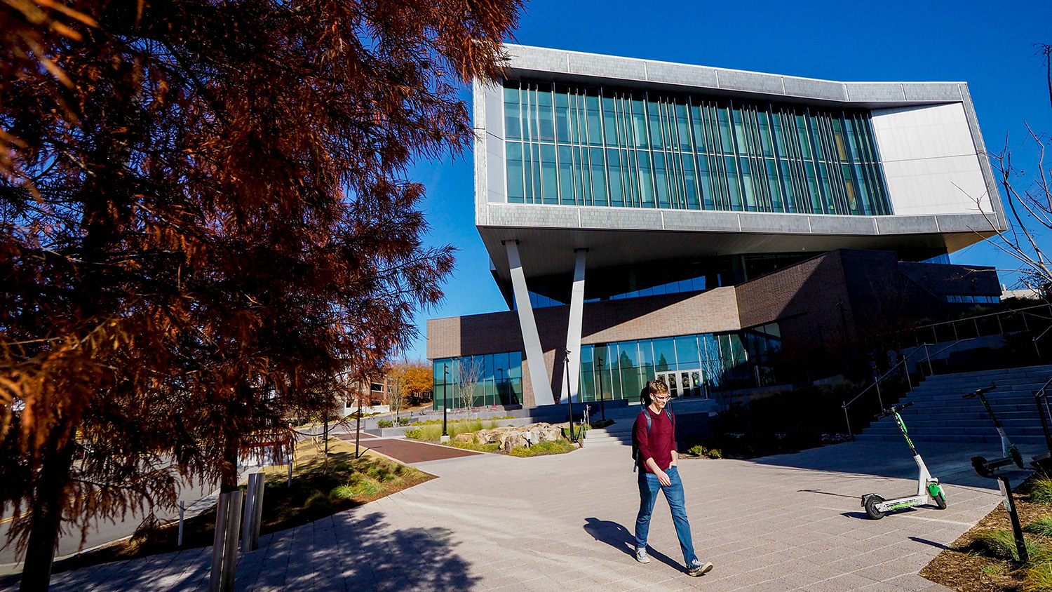 Student strolling along sidewalk during afternoon with the front of Fitts-Woolard Hall and clear blue sky in the background.