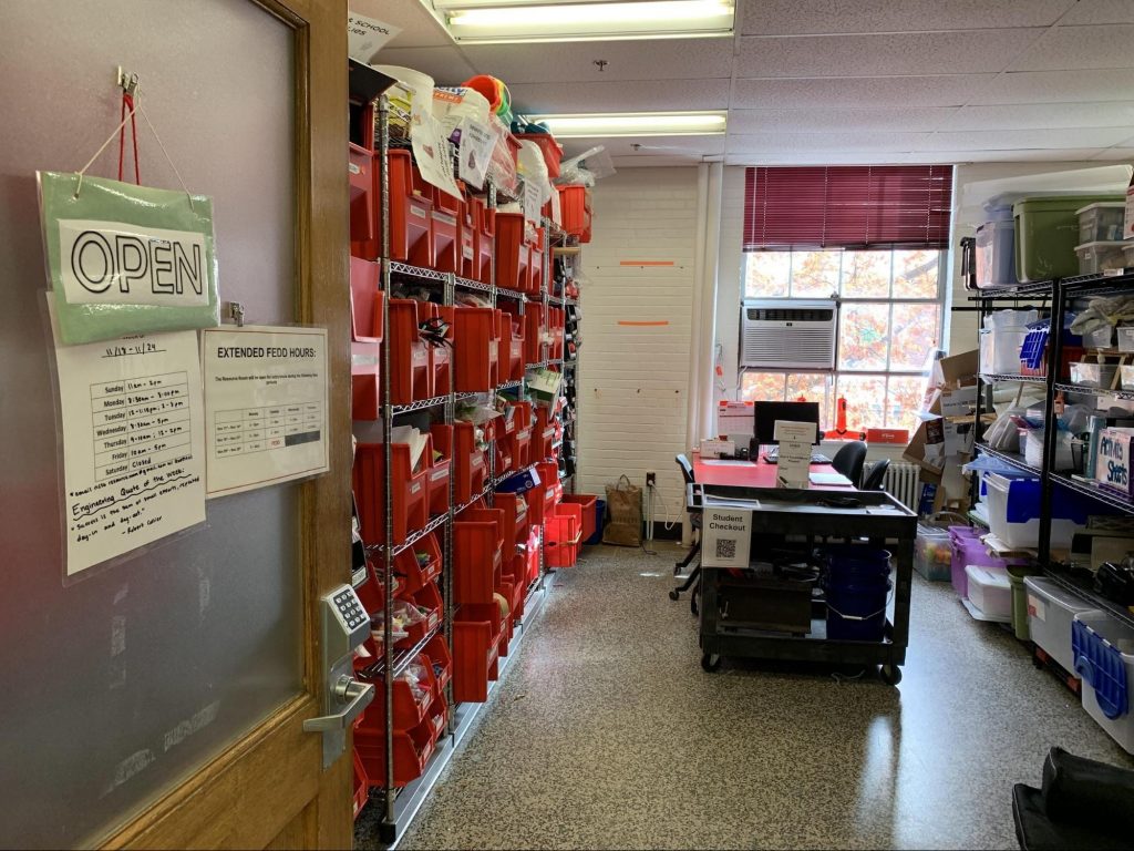 view of inside The Engineering Lab: on left is large chrome shelving with large red plastic containers; in the middle is a stationary desk, a mobile desk and to the right another shelving unit holding a variety of containers and supplies; there is a large window behind the desks.
