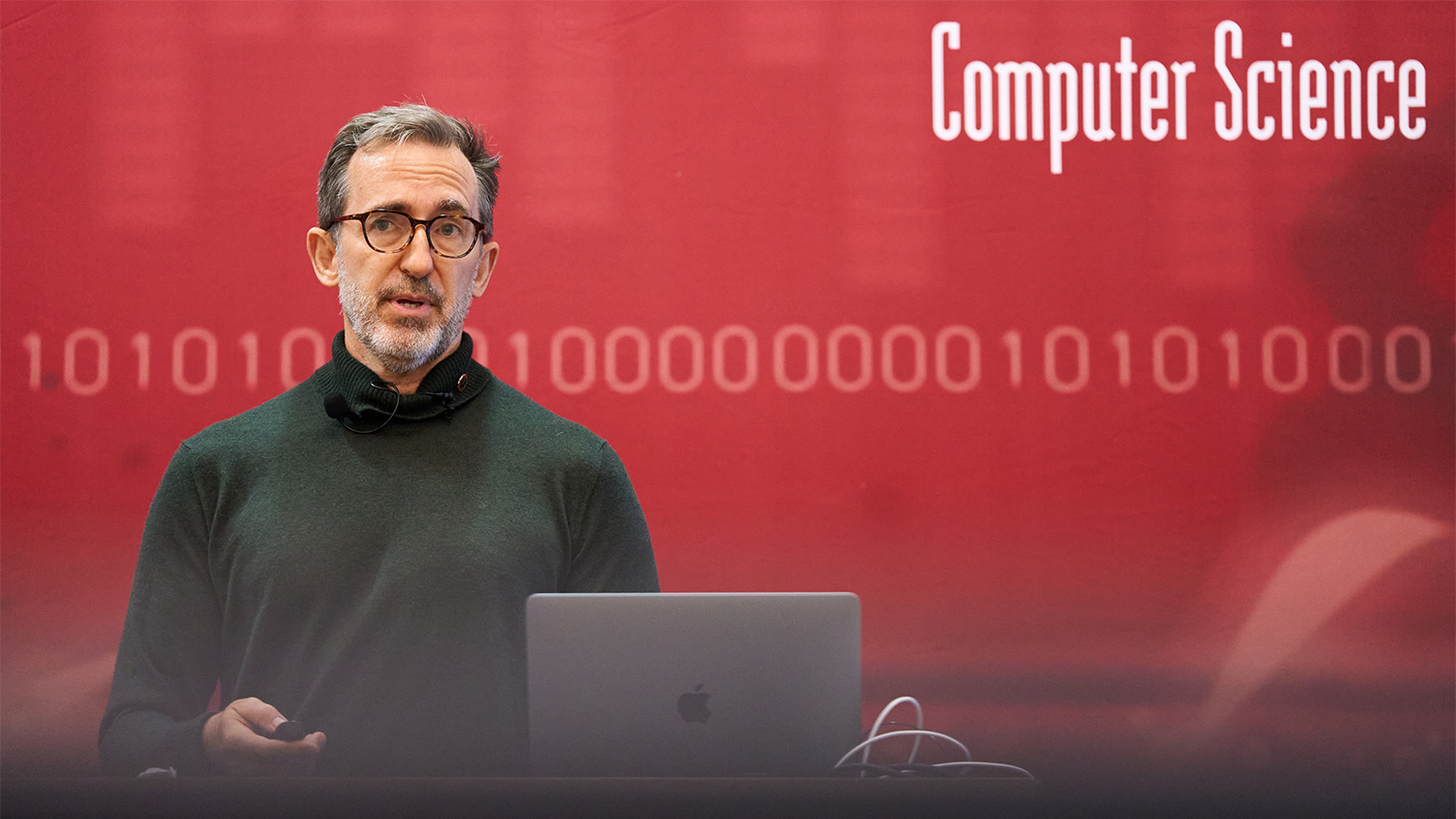 Drew Banks, standing behind an Apple laptop, speaks to a group which is outside of view of the frame.