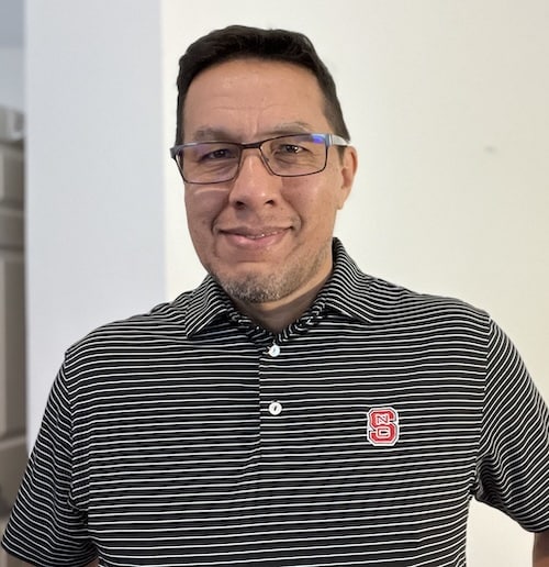 man with dark hair, wearing glasses and a dark gray polo shirt with the NC State logo attached