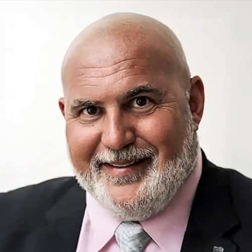 older man with bald head, white beard, wearing a dark suit jacket, pink shirt and grey necktie