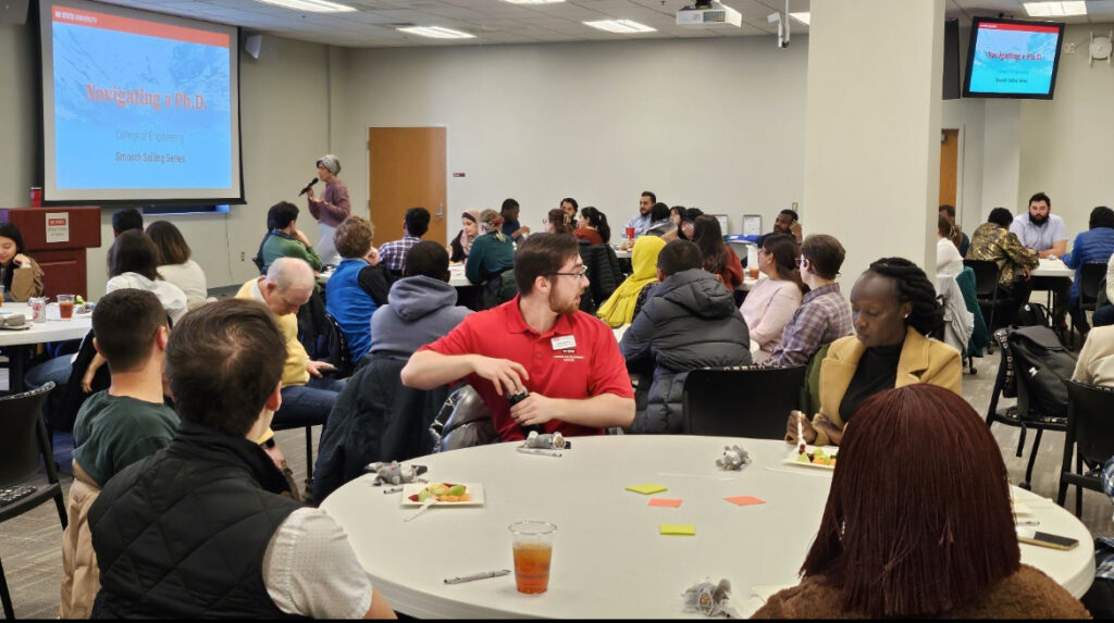 NC State graduate students all seated at round tables in a large conference room with off-white walls. A large screen is on the left and a smaller one in the upper right.