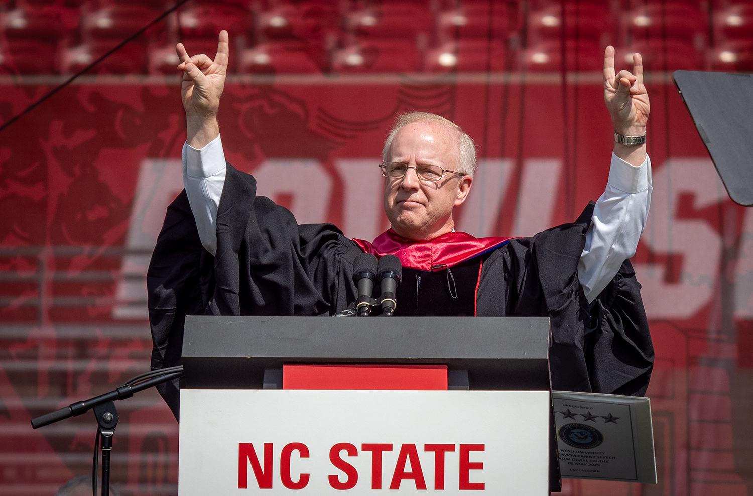 Daryl Caudle dressed in graduation robe give the NC State wolf sign while standing behind a podium