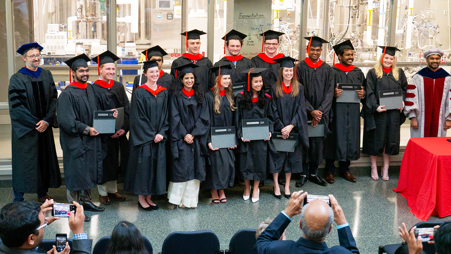 graduating students dressed in black gowns and caps, and red sashes pose as a group for photos