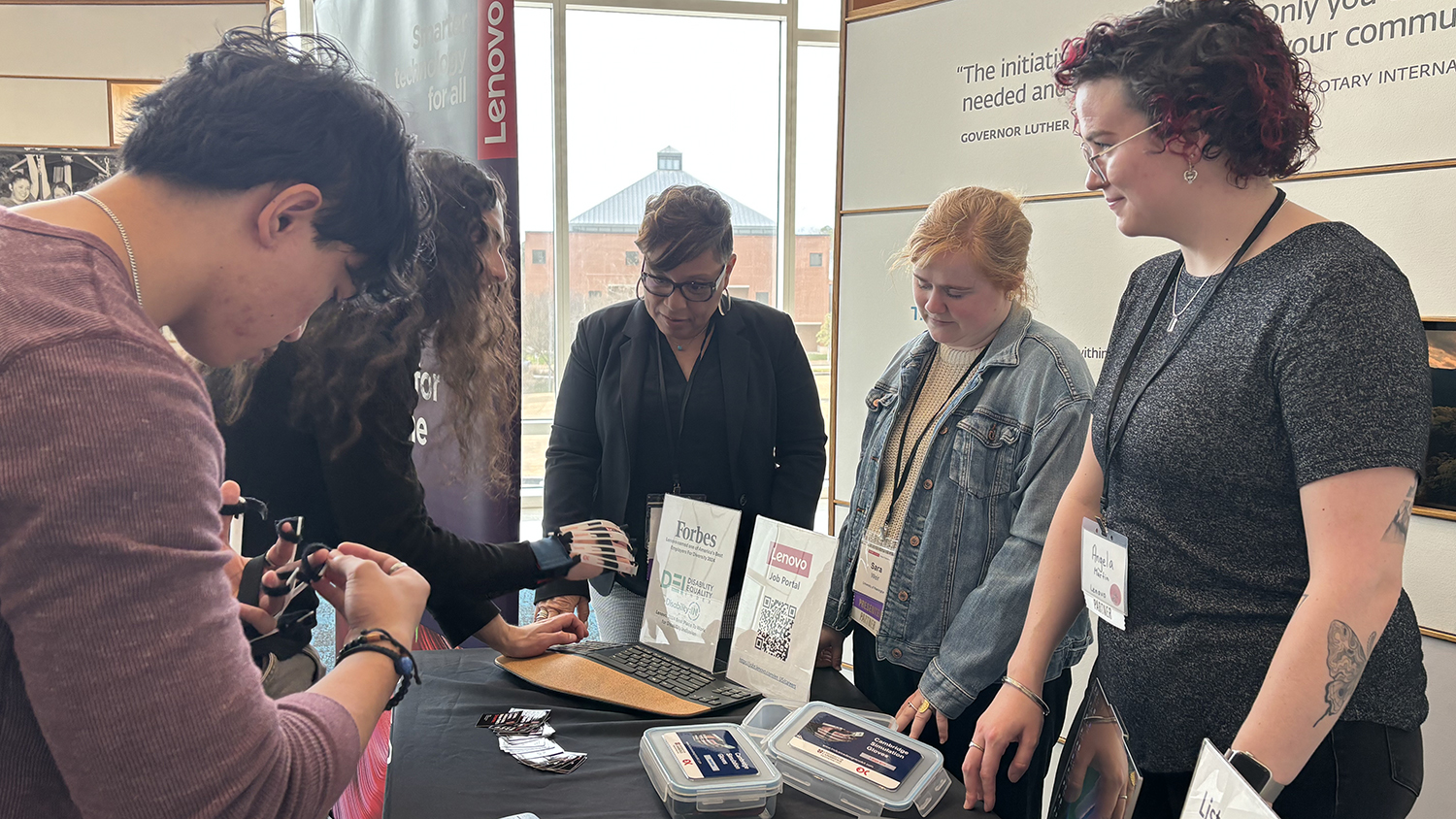 group of conference attendees gather around a table to watch a demonstration of an accessible device for use on a hand