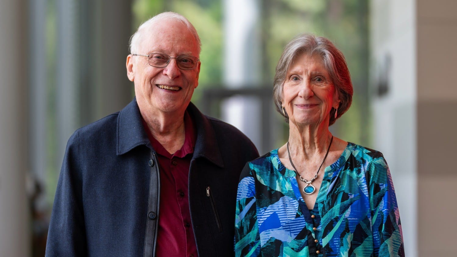 Smiling elderly couple indoors, wearing glasses and colorful attire, standing close together, natural light in background.